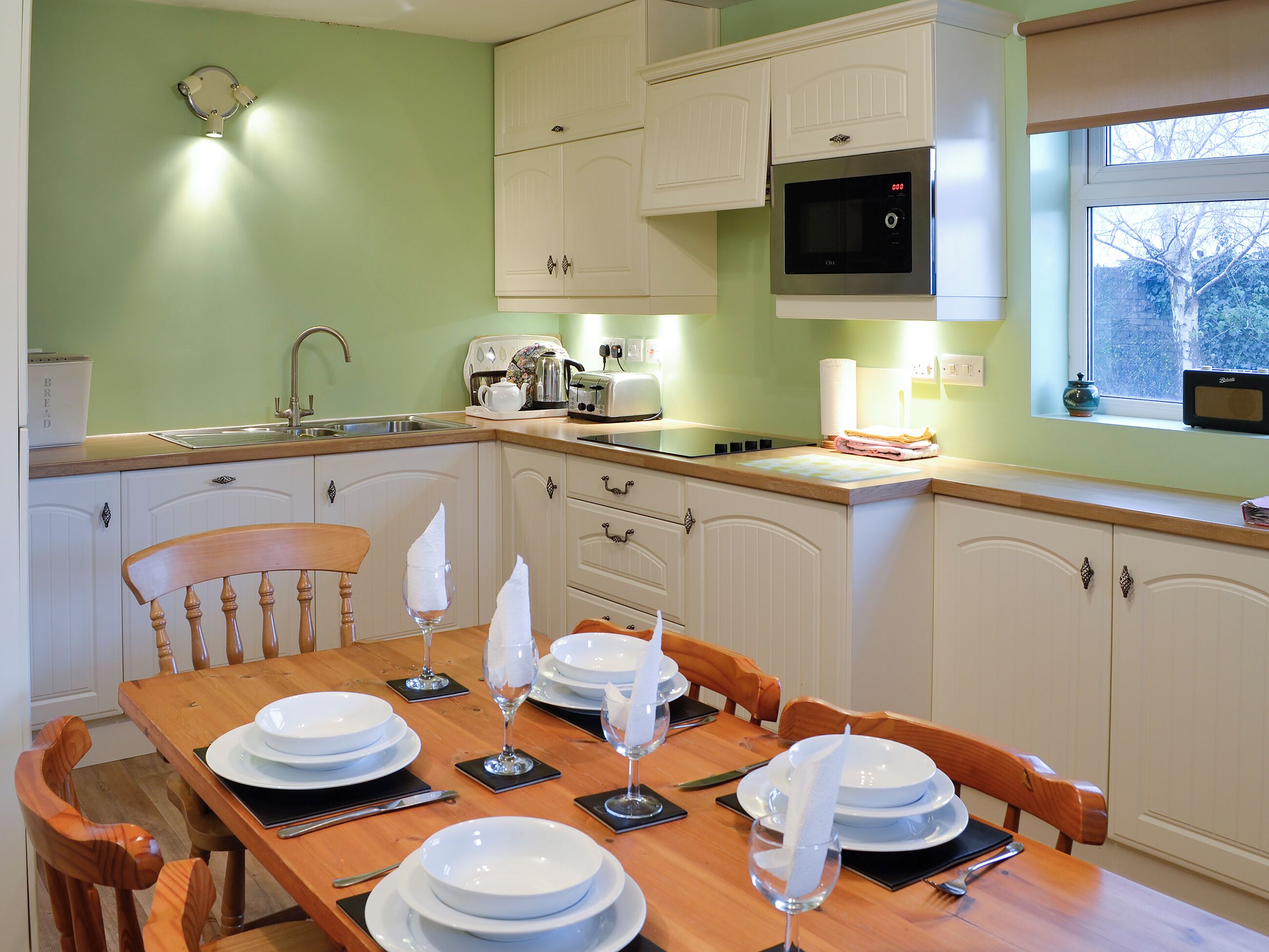 Una cocina con una mesa de comedor para cuatro, armarios blancos, un microondas y una ventana con vistas a árboles.