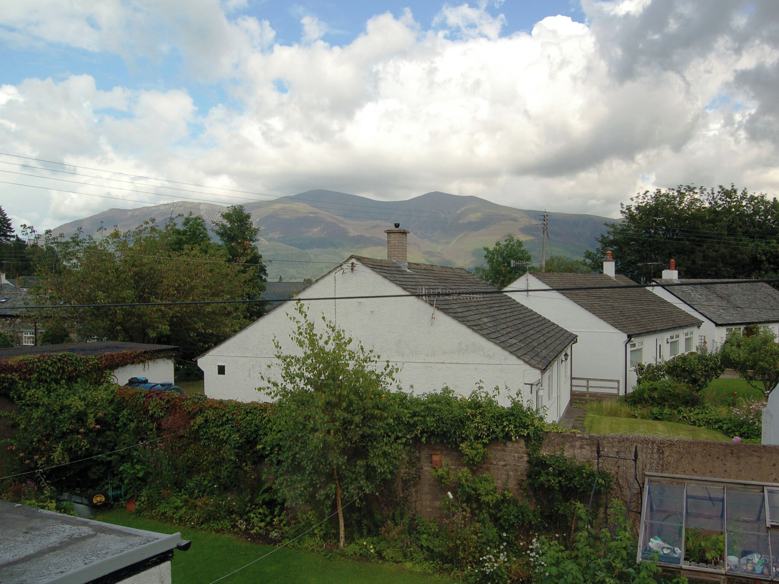 Ein ländliches Haus mit Schrägdach, umgeben von viel Grün und einem Berg im Hintergrund.