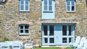 Un edificio de piedra con puerta y ventanas azules, mobiliario exterior con sillas y mesas, y un césped bien cuidado.