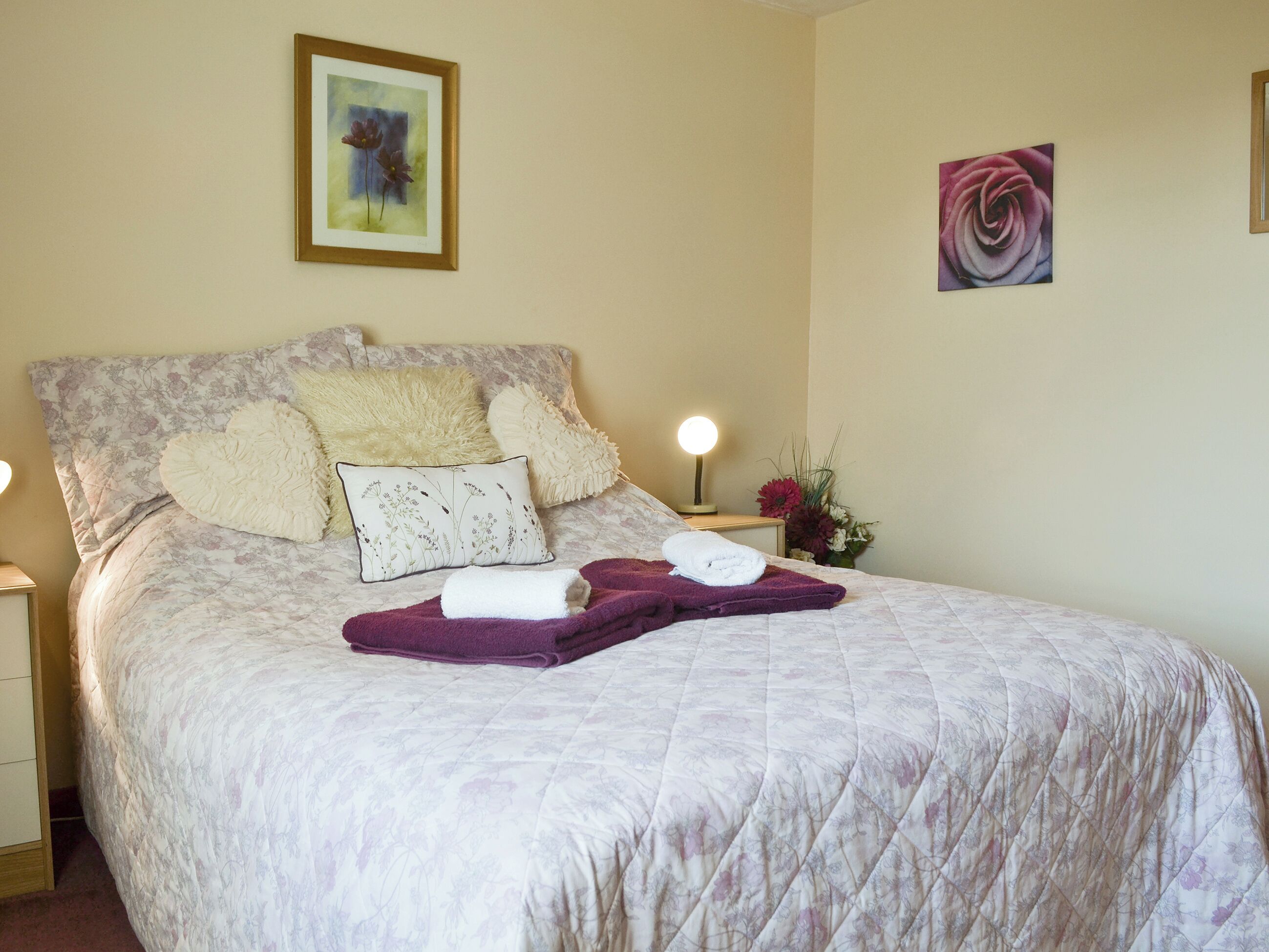 Un lit bien fait, avec une literie à motifs floraux, un plaid violet et deux serviettes. Deux lampes murales et un tableau floral encadré sont accrochés au mur, au-dessus du lit.