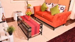 A living room with a red sofa, a small table with a red and white cloth, a white side table, a potted plant, and a vase with flowers. - Sherry Homes Zuri Studio (Nairobi)