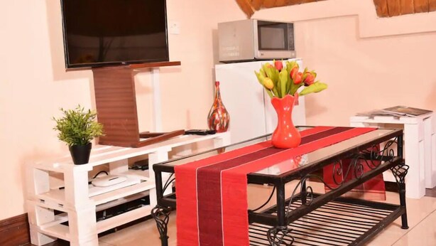 A living room with a flat-screen TV, a white entertainment unit, a red and black table, a small plant, and a vase with flowers. - Sherry Homes Zuri Studio (Nairobi)