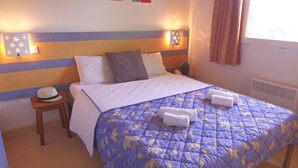 A neatly made bed with a blue and white quilt, two pillows, and two folded towels on the bed. There is a small wooden nightstand with a hat on it next to the bed. - VVF La Salvetat sur Agoût (La Salvetat-sur-Agout)