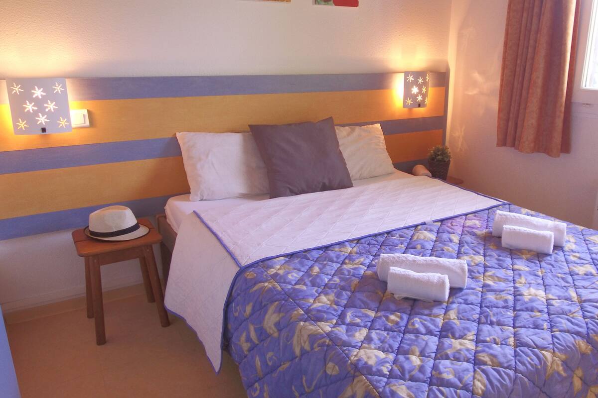 Un lit bien fait, recouvert d’une couverture bleue et blanche, avec deux oreillers et deux serviettes pliées posées dessus. Une petite table de chevet en bois, sur laquelle repose un chapeau, se trouve à côté du lit.