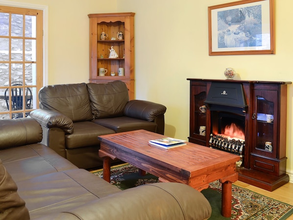 A living room with a brown leather sofa, a wooden coffee table, a fireplace, and a wooden cabinet with shelves. - The Stables at Old Vicarage Cottage (Betws-Yn-Rhos)