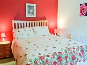 A bedroom with a red wall, a wooden headboard, floral bedding, two bedside lamps, and a framed picture above the bed.