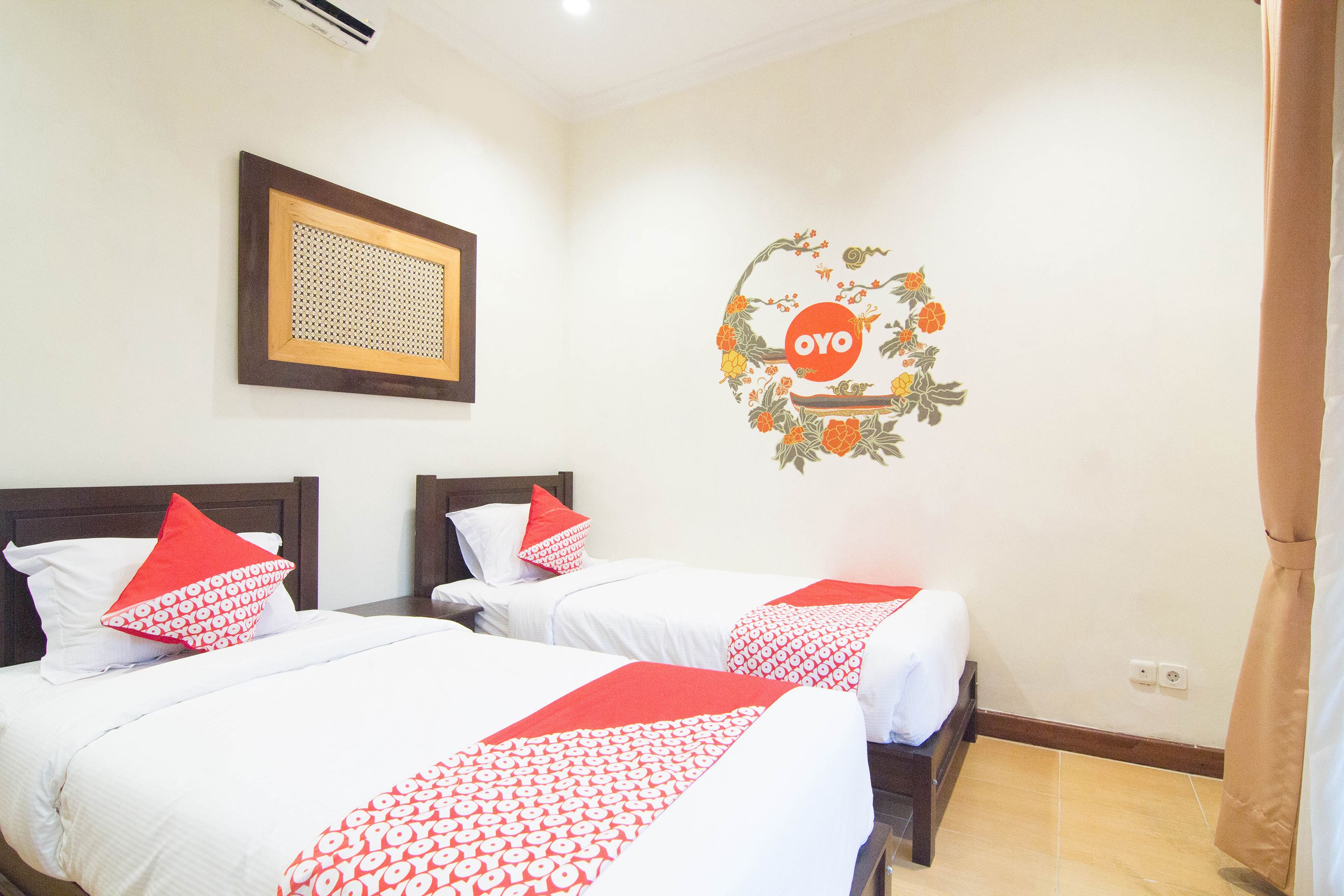 A hotel room with two beds, each with white bedding and red accents, a framed artwork on the wall, and a logo with the word "OYO" on the wall.