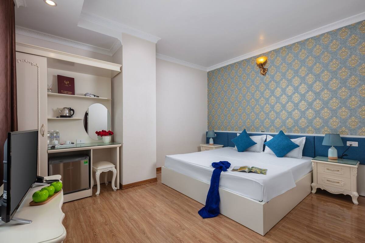 A hotel room with a bed, bedside tables, a desk with a computer, a small fridge, and a wardrobe.
