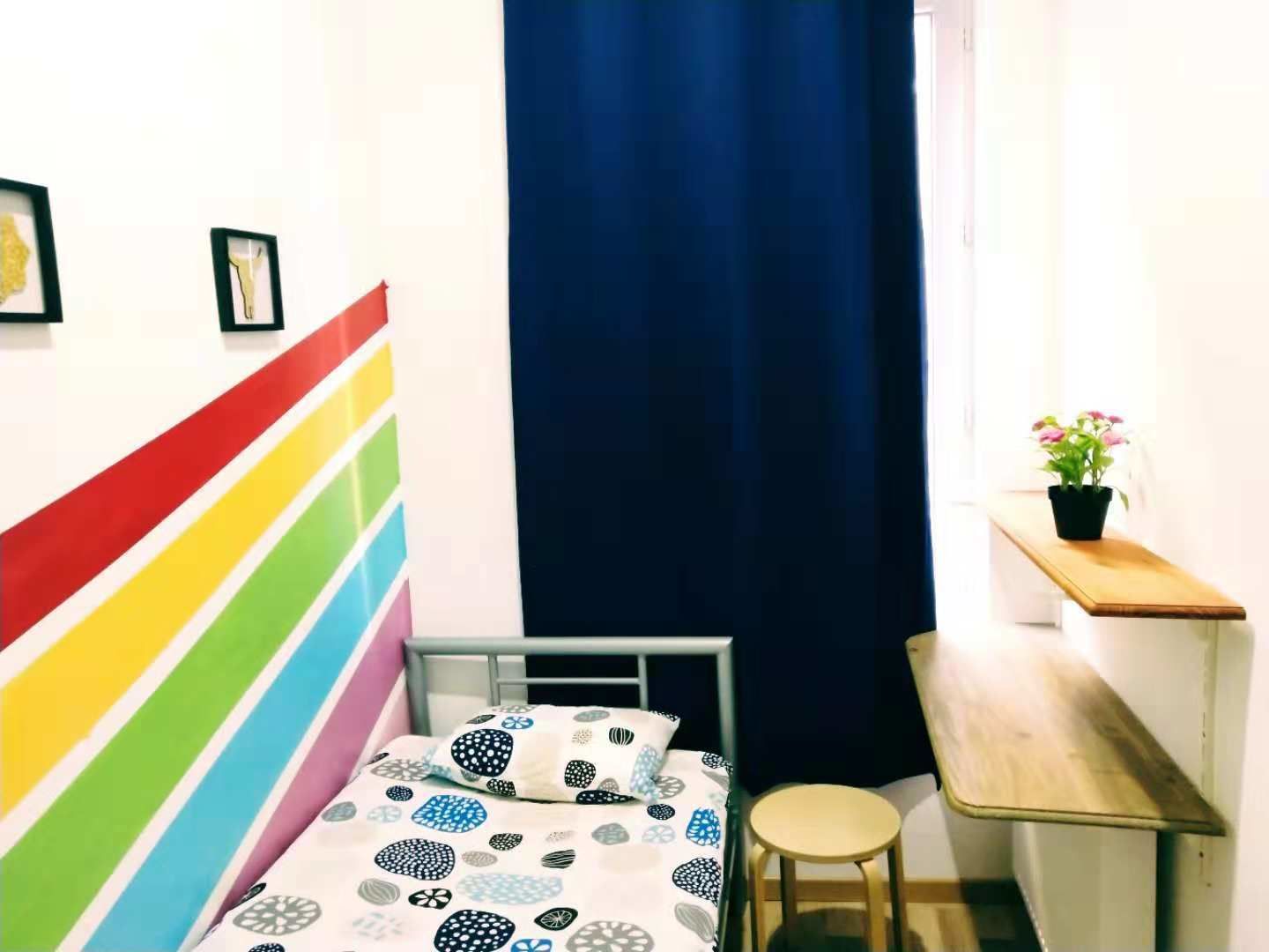 Pieni huone, jossa on yksi sänky, värikäs raidallinen pääty ja työpöytä jakkaralla sekä ruukussa oleva kasvi.
