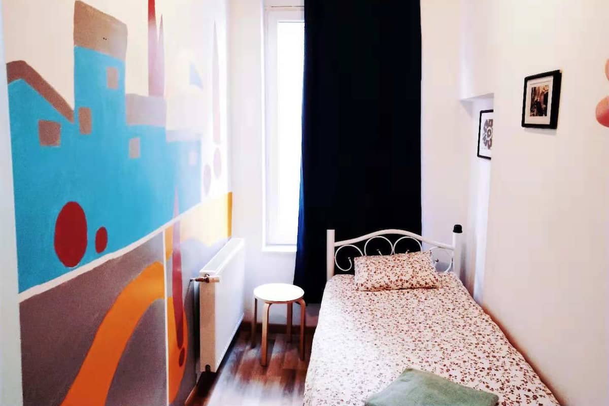 Ein kleines Schlafzimmer mit Bett, Heizkörper, einem Fenster mit Vorhängen und einer Wand mit abstrakter Kunst.