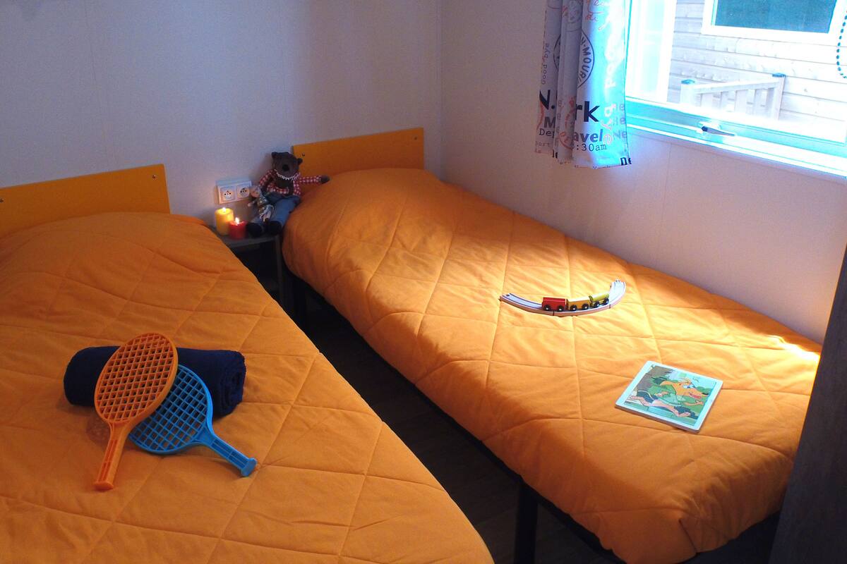 A room with two beds, each with an orange bedspread. There is a small table with a candle and a teddy bear. A window with curtains is visible, and a toy train is on the right bed.