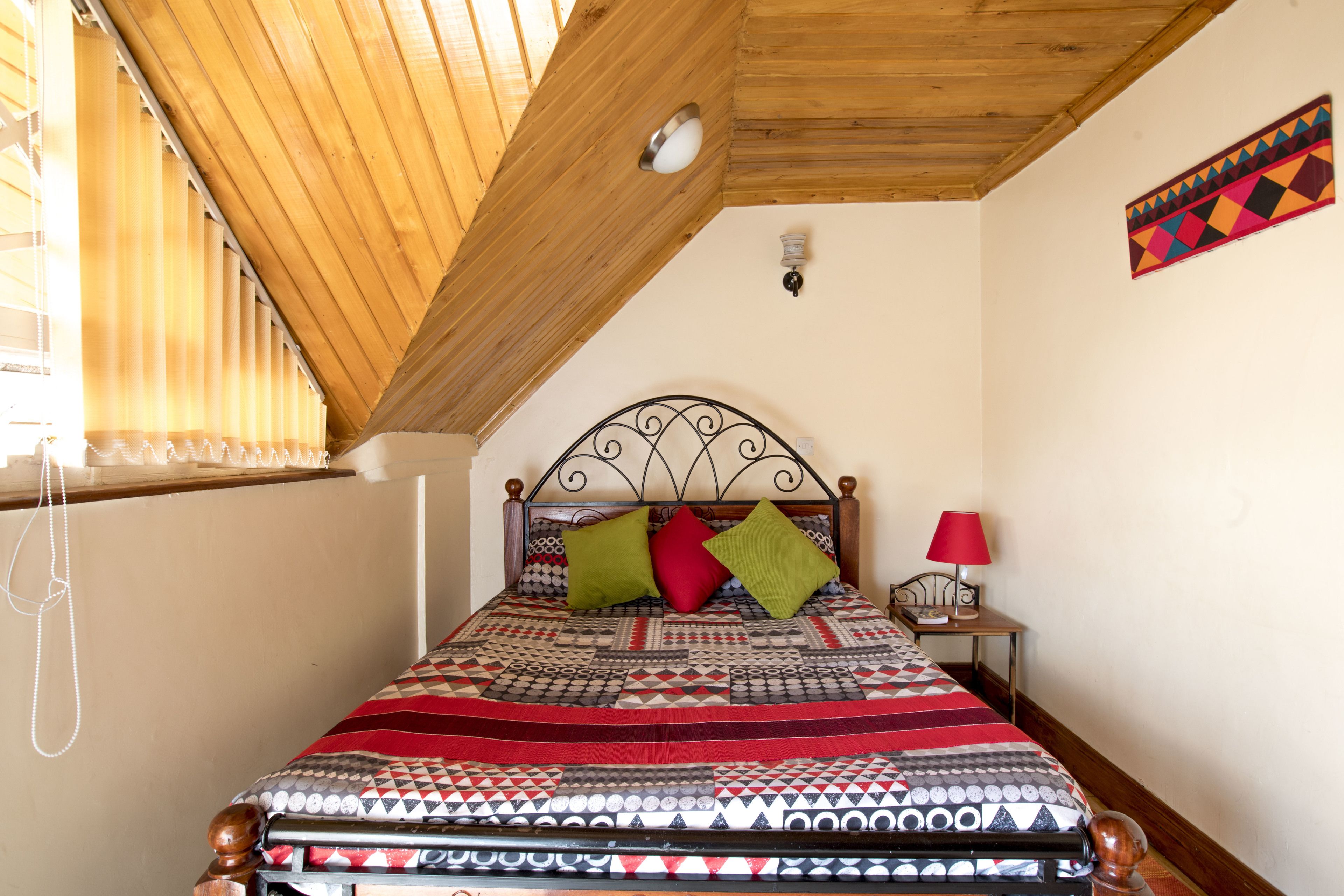Une chambre avec un plafond en pente, un lit recouvert d’une couvre-lit à motifs, deux oreillers, une table de chevet avec une lampe et un luminaire fixé au mur.
