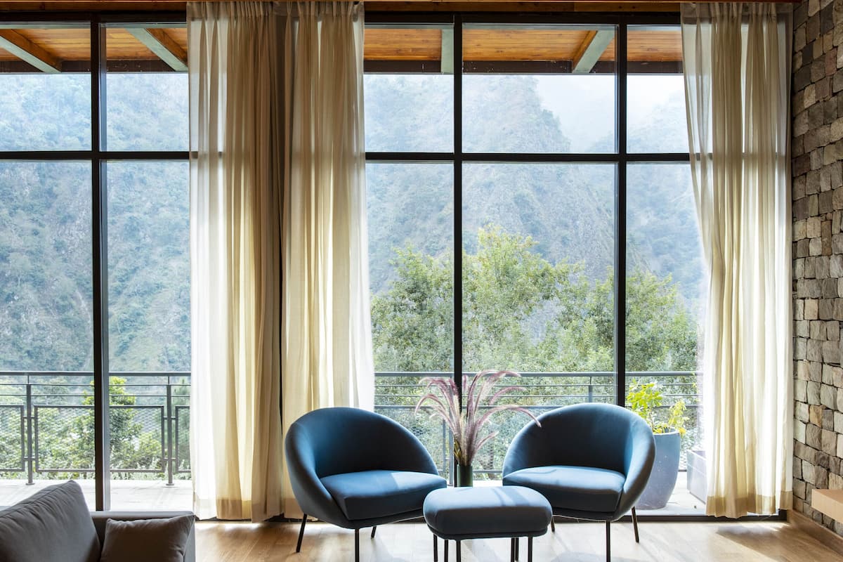 A room with large windows, a wooden ceiling, and a view of a mountainous landscape. There are two blue chairs and a wooden floor.