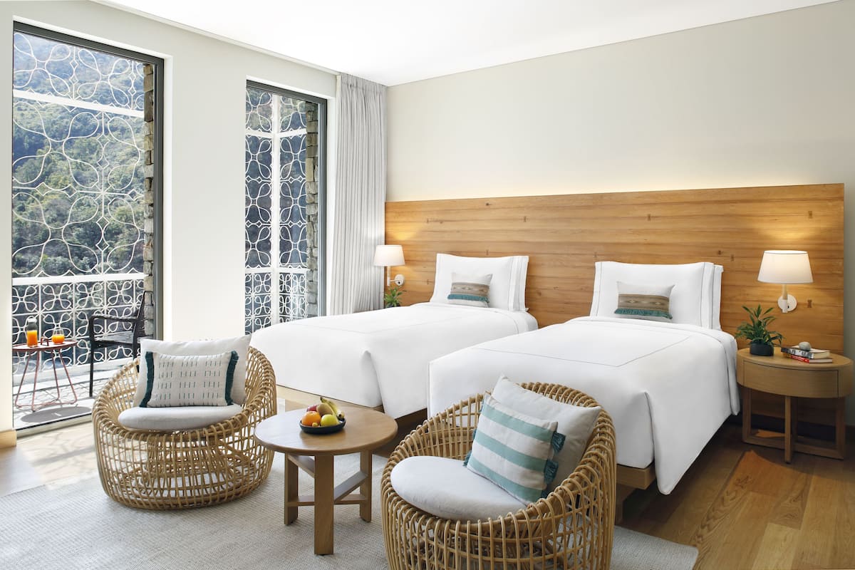 A hotel room with two beds, wicker chairs, a small table with fruit, and a balcony with a view of greenery.