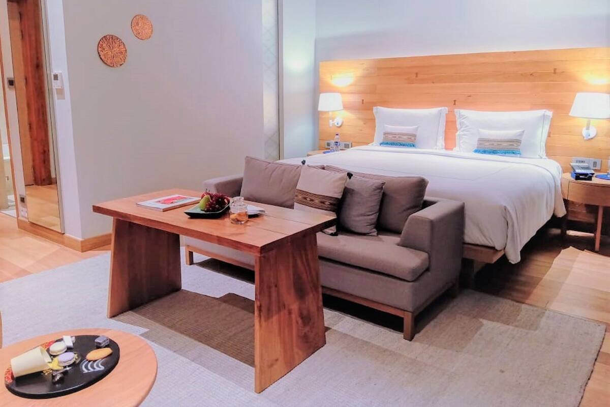 A hotel room with a bed, a wooden table, a sofa, and a small round table with snacks.