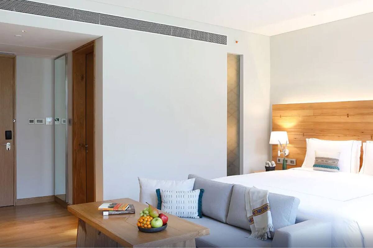 A modern hotel room with a large bed, a wooden headboard, a bedside table with a lamp, and a fruit bowl on a small table.