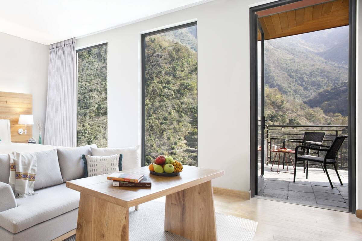 A modern hotel room with a large bed, a sofa, a wooden coffee table with books, and a balcony with a mountain view.