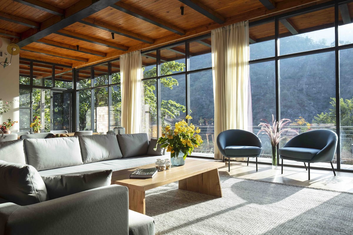 A spacious living room with a wooden ceiling, large windows, a sofa, two armchairs, a coffee table, and a vase with flowers.