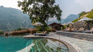 A pool area with lounge chairs and umbrellas, surrounded by stone walls and vegetation, with mountains in the background. - Taj Rishikesh Resort & Spa, Uttarakhand (Narendranagar)
