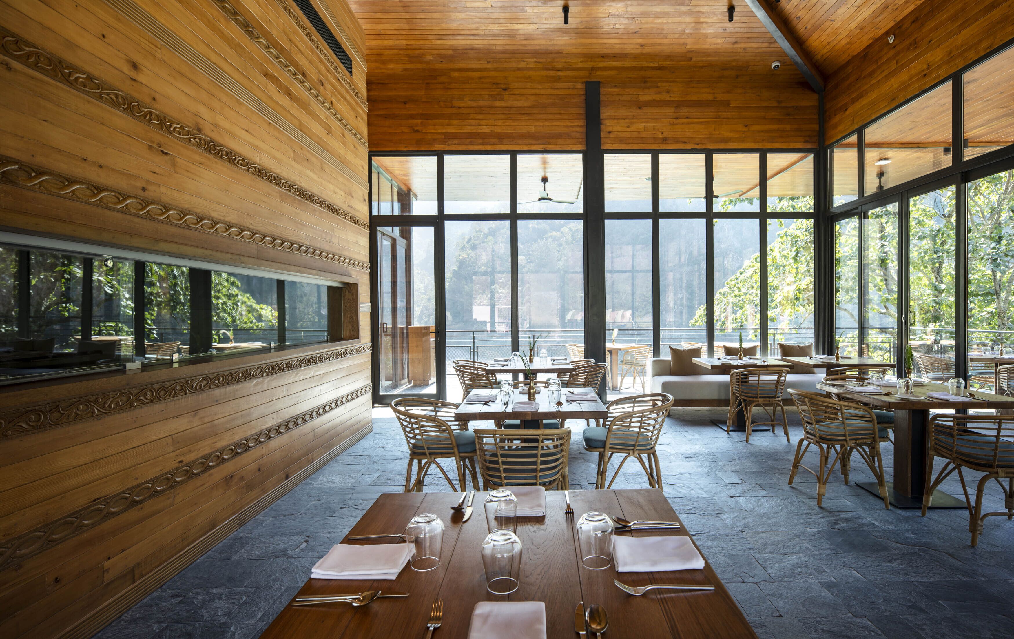 A restaurant with wooden walls and large windows overlooking a body of water. Tables are set with cutlery and glasses.