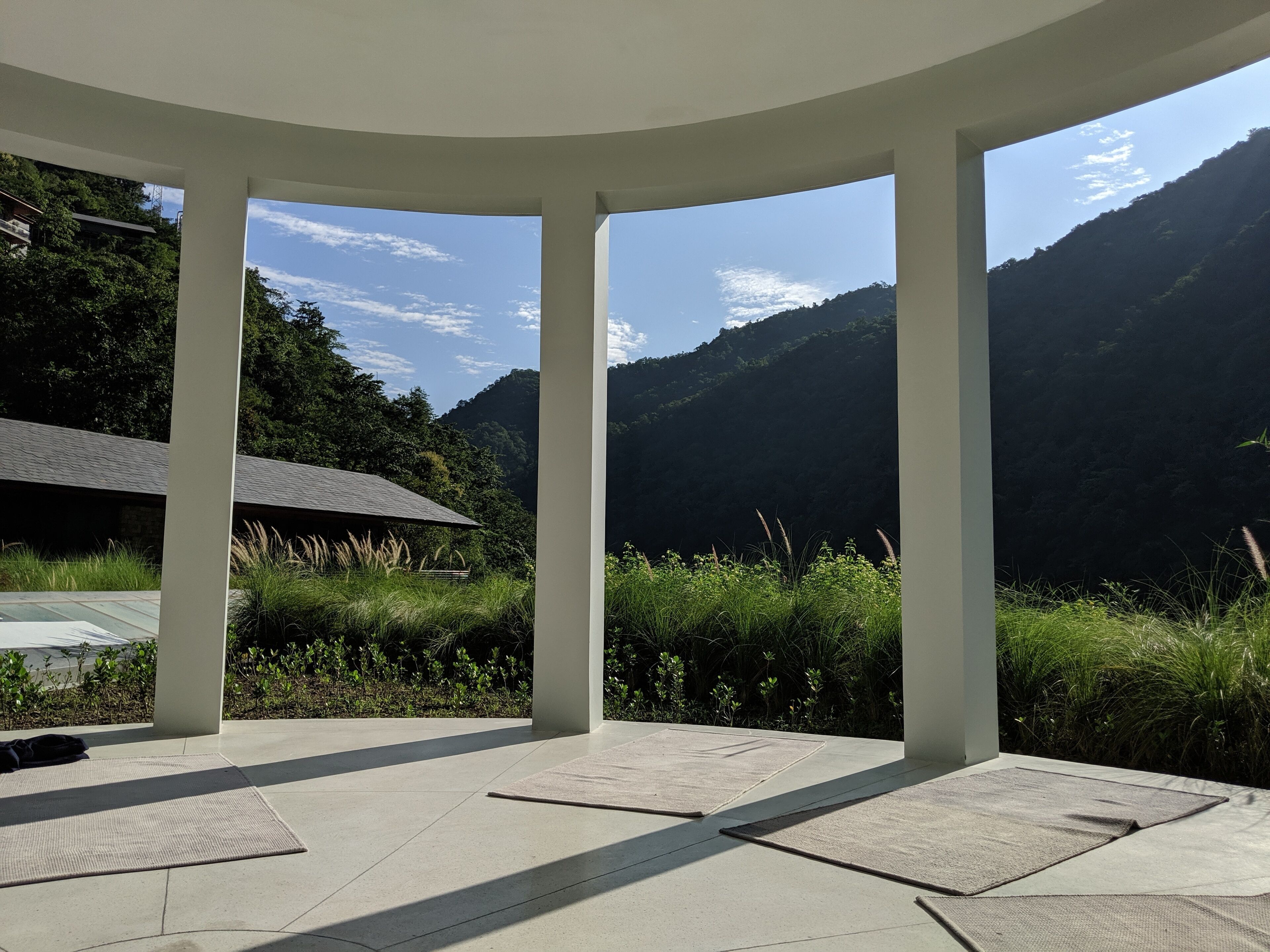 A covered outdoor area with a view of mountains, a building with a sloped roof, and a paved floor with mats.