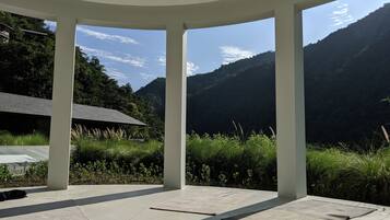 A covered outdoor area with a view of mountains, a building with a sloped roof, and a paved floor with mats.