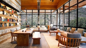 A spacious reading area with a wooden table, benches, and a sofa, surrounded by large windows offering a view of a mountainous landscape. - Taj Rishikesh Resort & Spa, Uttarakhand (Narendranagar)