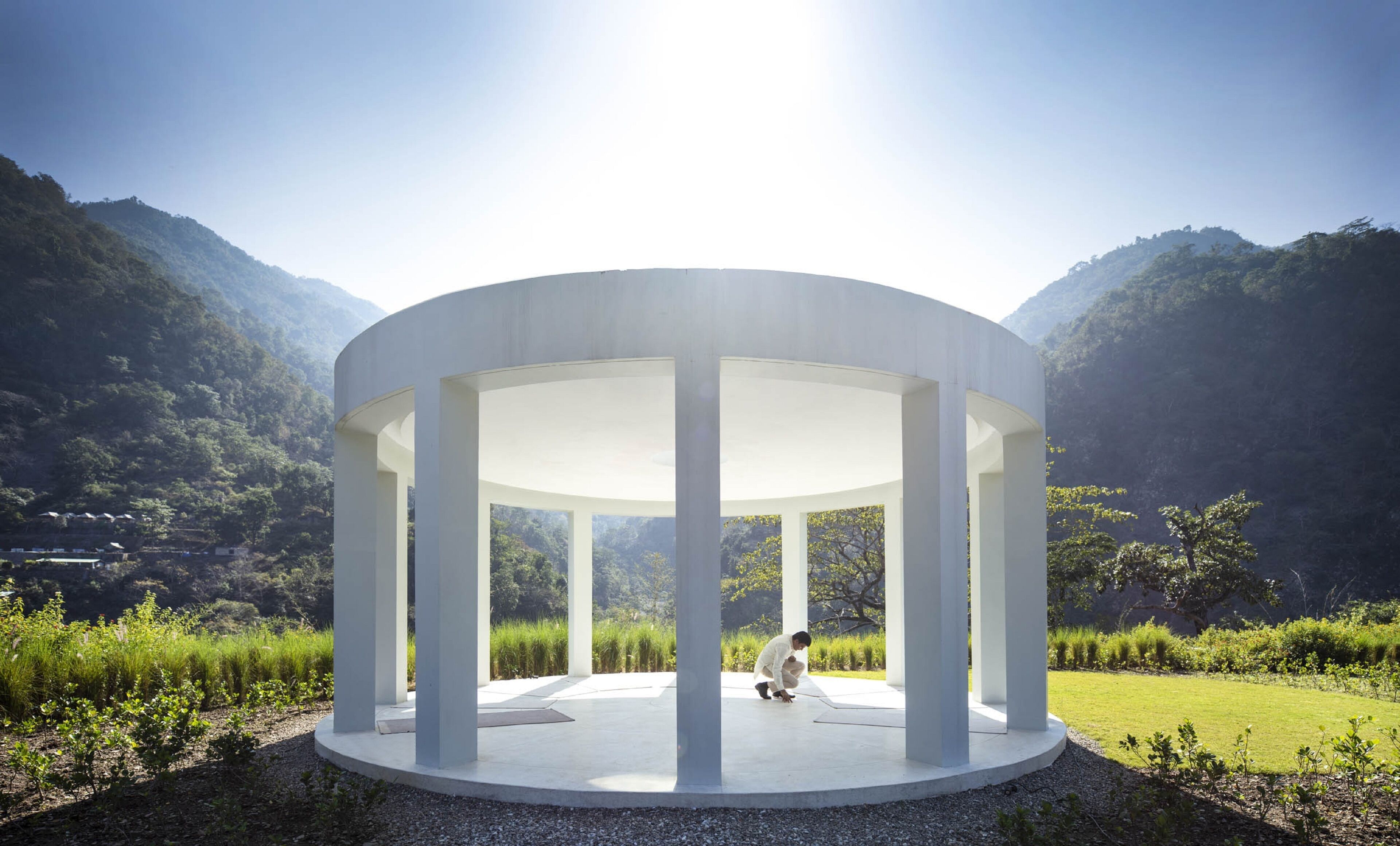 A circular pavilion with a central open space, surrounded by a gravel path and greenery, set against a mountainous backdrop.
