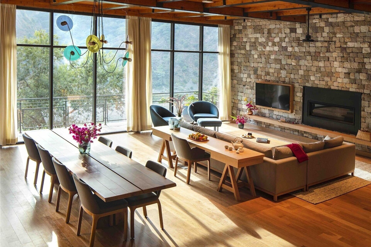 A spacious living area with a stone wall, wooden beams, a fireplace, and large windows.