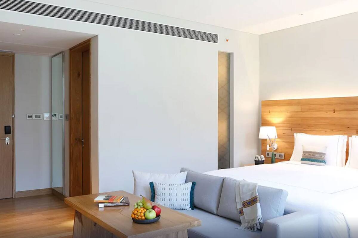 A hotel room with a bed, a bedside table with fruit, a sofa, a wooden headboard, and a nightstand with a lamp.