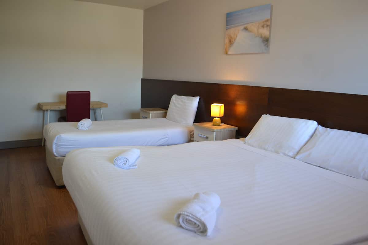 A hotel room with two beds, a nightstand with a lamp, a red chair, and a painting on the wall.