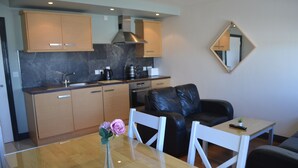 A modern kitchen with wooden cabinets, a marble backsplash, and a stainless steel range hood. There is a dining area with a wooden table and chairs, and a black leather sofa. A small potted plant is on the table.