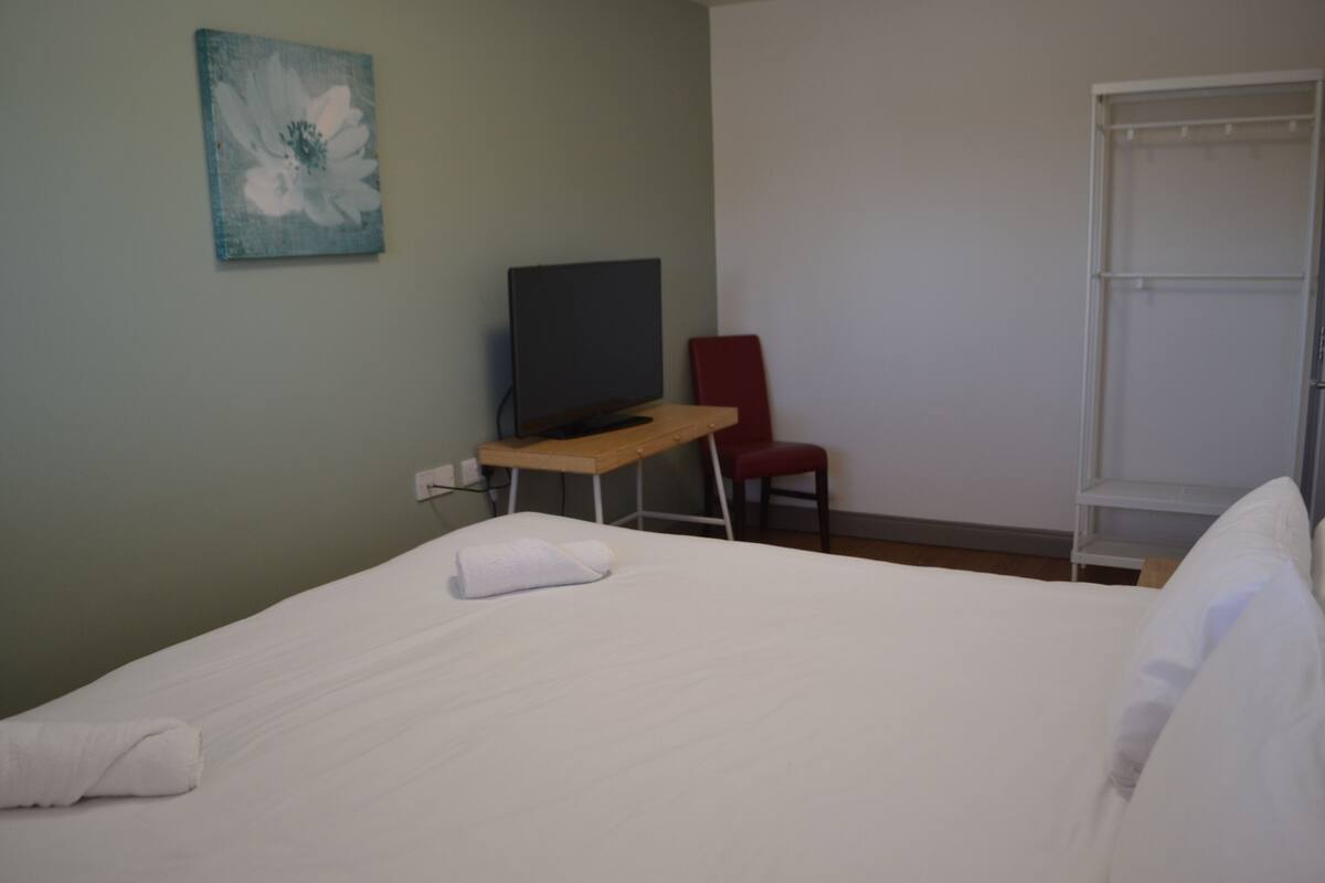 A hotel room with a bed, a desk with a TV, a red chair, and a wardrobe.