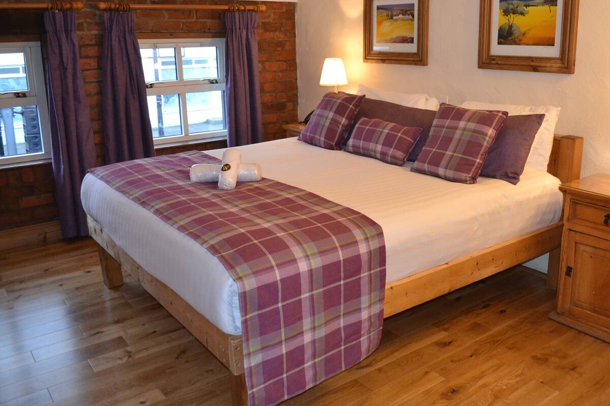 A wooden bed with a plaid bedspread and pillows, a wooden nightstand, a window with curtains, and a lamp.