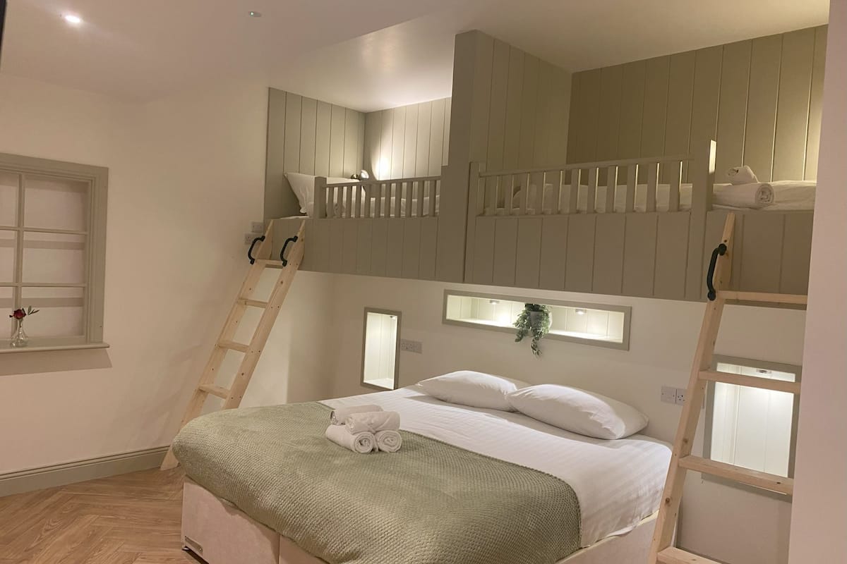 A bunk bed room with a ladder, a window with a plant, and a bed with a green blanket.