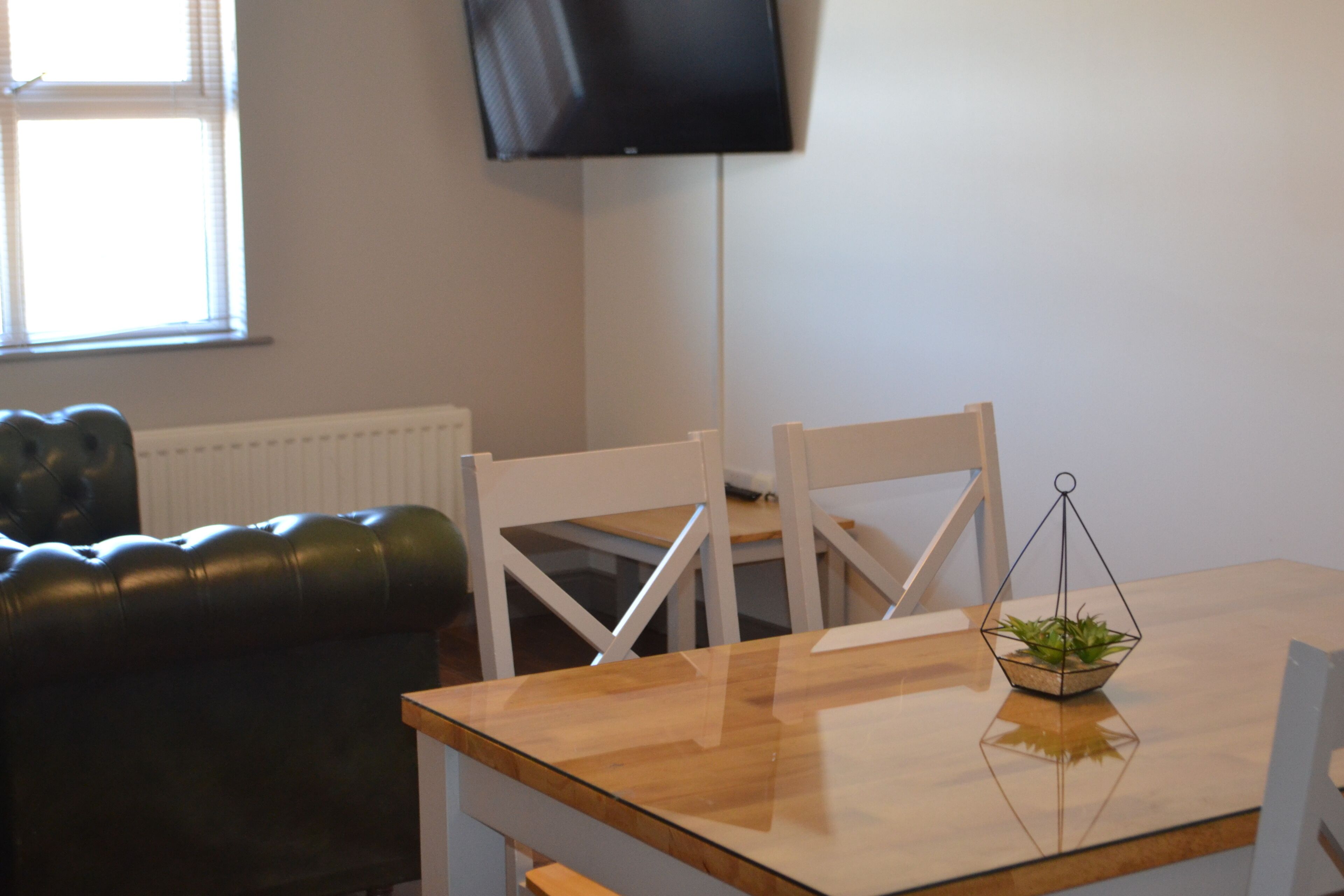 A room with a wooden table, two white chairs, a black leather couch, a black TV on the wall, and a small plant on the table.