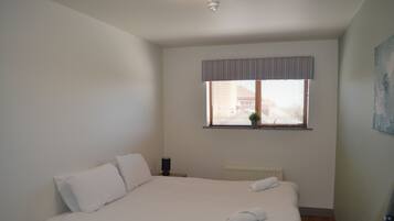 A bedroom with a bed, pillows, a nightstand, a window with blinds, a radiator, and a painting on the wall.