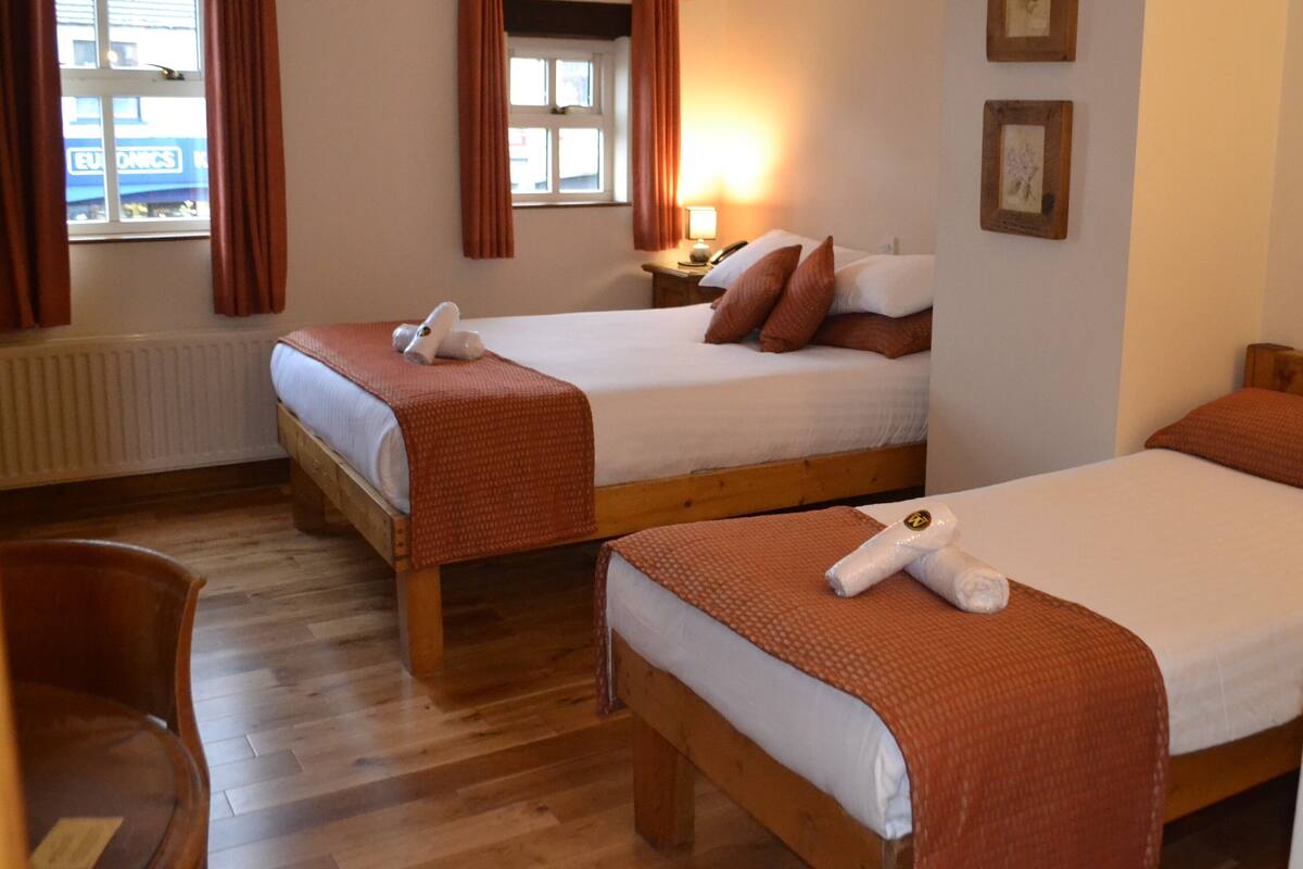 A hotel room with two single beds, wooden flooring, a radiator, a window with curtains, a bedside table with a lamp, and a chair.