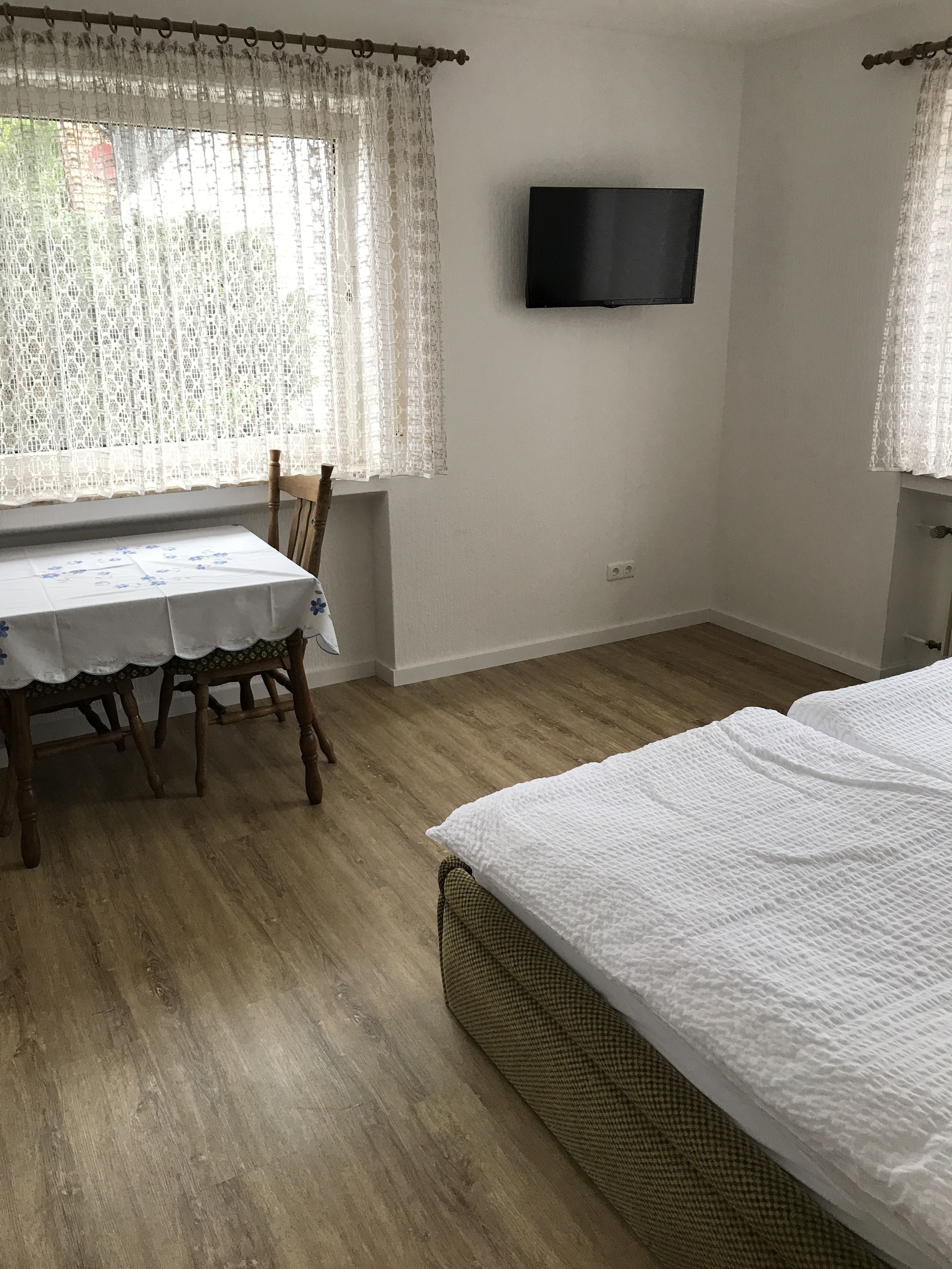 A room with a bed, a table with chairs, a television mounted on the wall, and lace curtains on the windows.