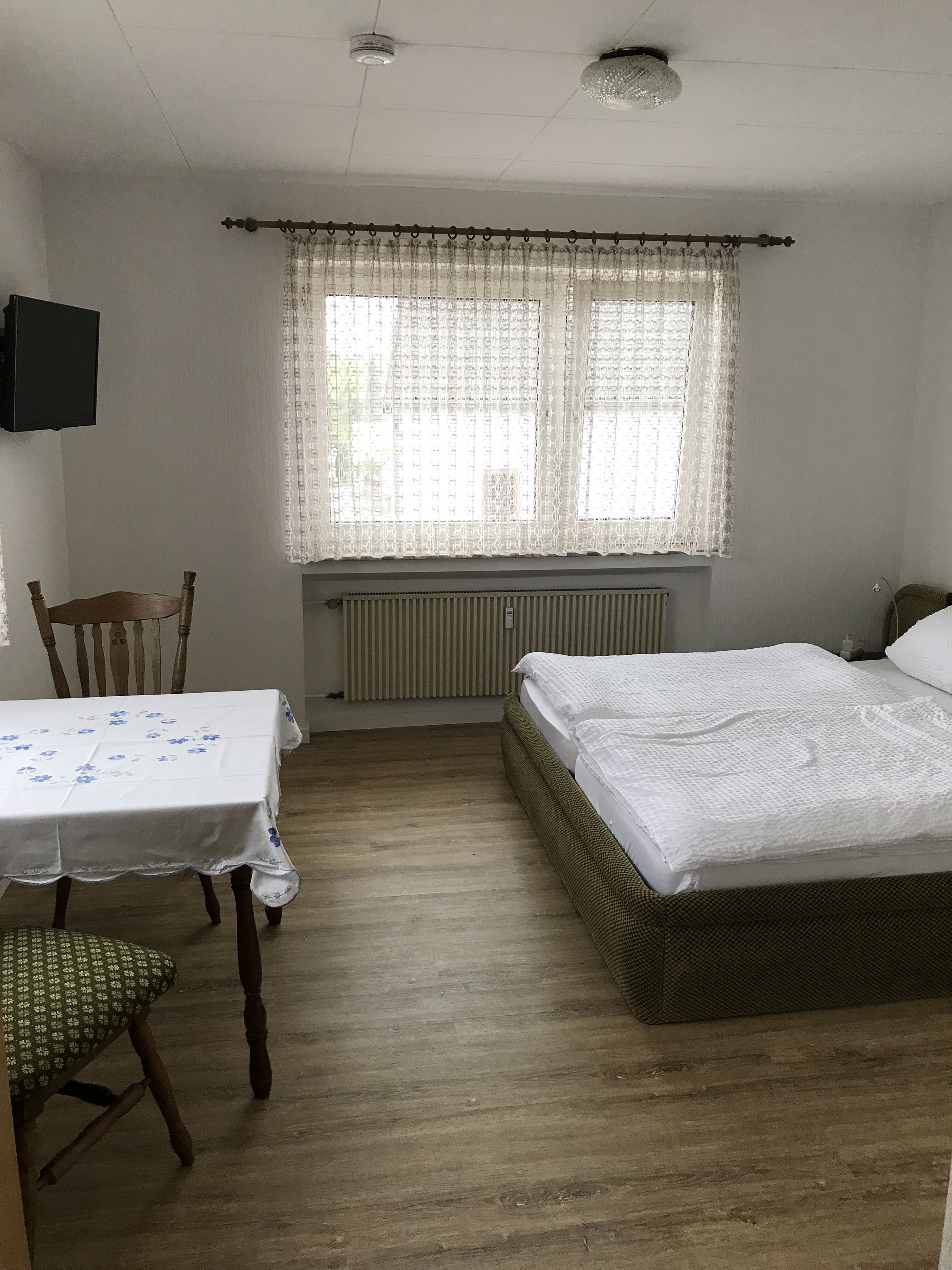 Une chambre à coucher avec un lit, une table, une chaise, un radiateur et une fenêtre avec des rideaux.