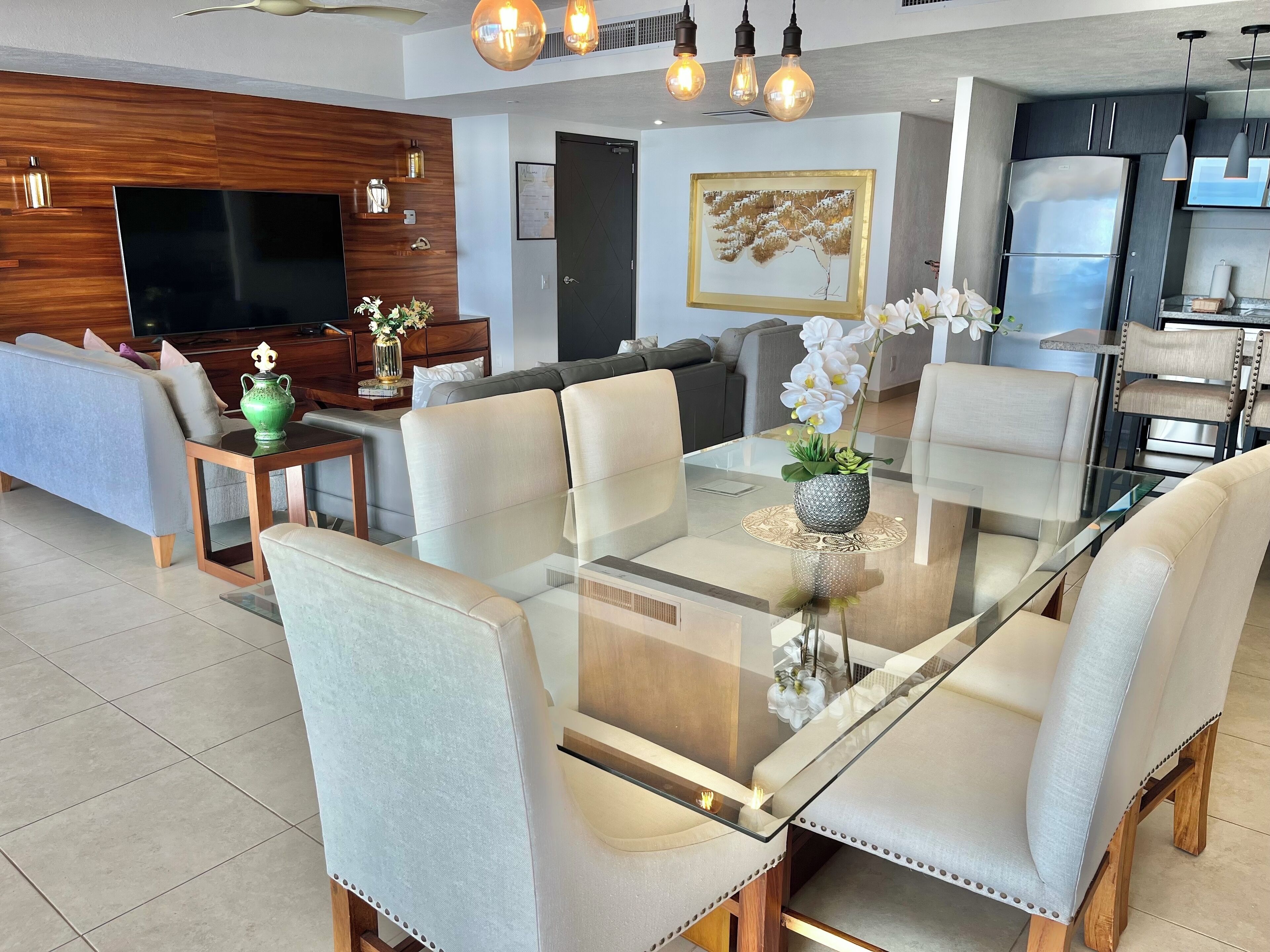 A modern living room with a glass dining table, comfortable seating, a flat-screen TV, and decorative elements.