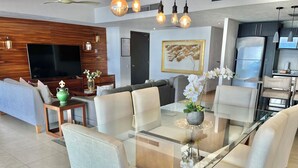 A modern living room with a glass dining table, comfortable seating, a flat-screen TV, and decorative elements. - 17th-Floor Beachfront Grand Venetian Condo ☆Panoramic Bay Views & Infinity Pools (Puerto Vallarta)