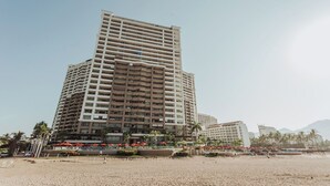 A multi-story hotel with a modern design, featuring balconies on each floor, located near a beach with palm trees and umbrellas. - 17th-Floor Beachfront Grand Venetian Condo ☆Panoramic Bay Views & Infinity Pools (Puerto Vallarta)