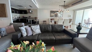 A modern living room with a black leather sofa, a dining area with a glass table, and a kitchen in the background. - 17th-Floor Beachfront Grand Venetian Condo ☆Panoramic Bay Views & Infinity Pools (Puerto Vallarta)
