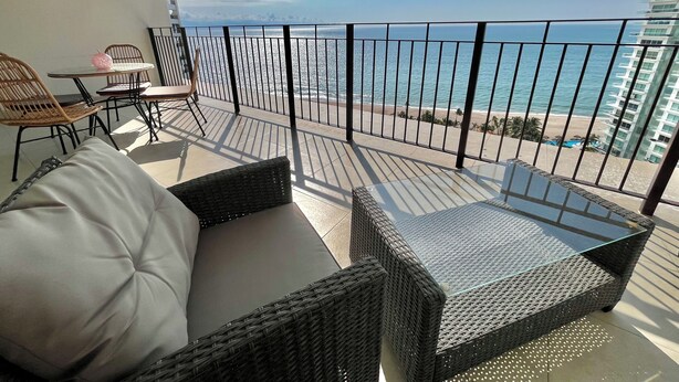 A balcony with wicker furniture, a glass table, and a view of the beach and ocean. - 17th-Floor Beachfront Grand Venetian Condo ☆Panoramic Bay Views & Infinity Pools (Puerto Vallarta)