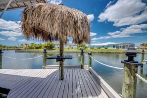A wooden dock with a thatched roof, a rope barrier, and a view of a building across the water. - Heated Pool | Gulf Access Canal | Dock | BBQ (Cape Coral)