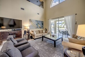 Un amplio salón con un televisor de pantalla plana grande, un sofá color beige, un sofá modular gris, una mesa de centro de vidrio y vistas al exterior a través de ventanas grandes.
