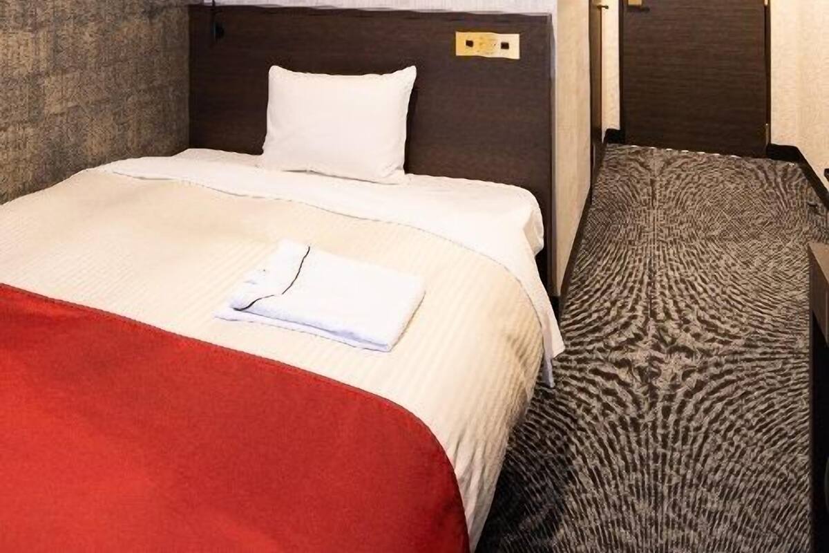 A hotel room with a bed, a red blanket, a white pillow, a headboard, a wall with a textured pattern, a door, and a carpet with a circular design.
