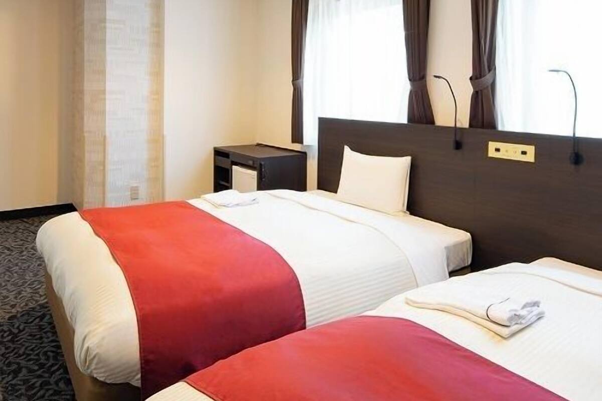 A hotel room with two beds, a bedside table, a lamp, and a window with curtains.