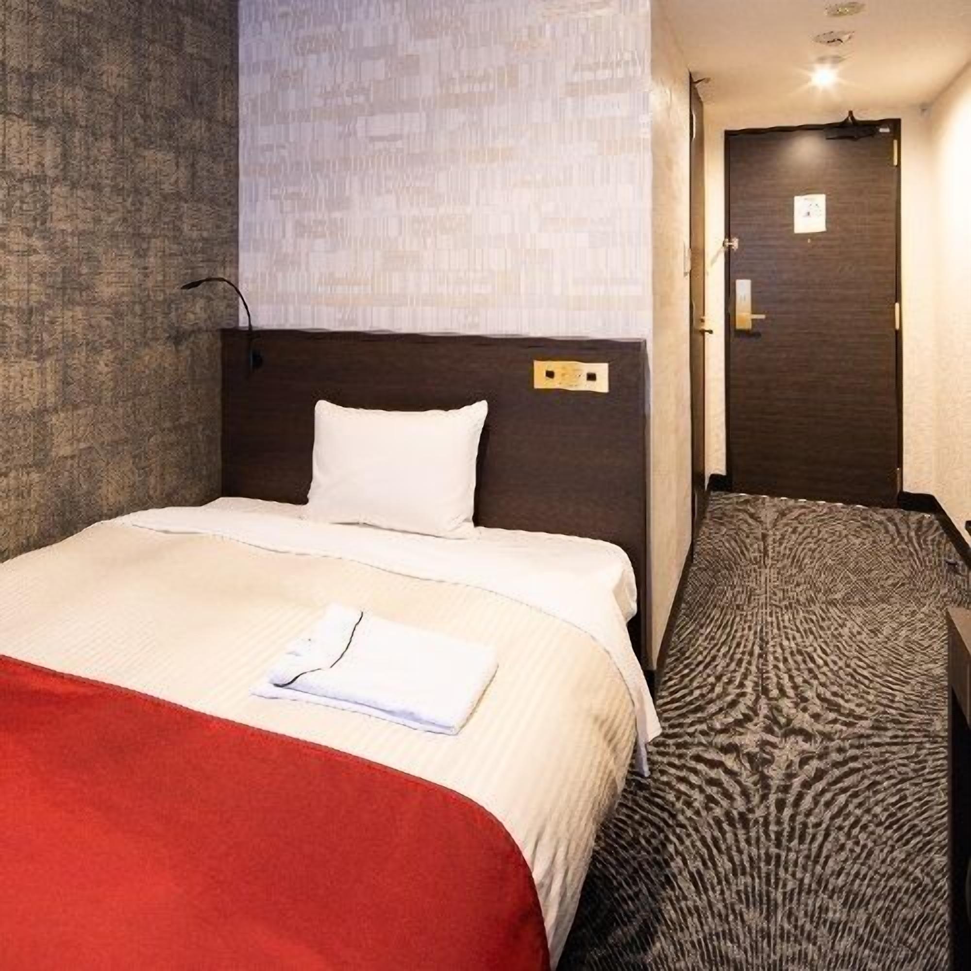 A hotel room with a bed, a red blanket, a white pillow, a headboard, a wall with a textured pattern, a door, and a carpet with a circular design.